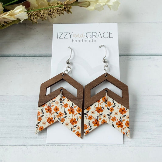 A pair of geometric drop earrings displayed on a white card labeled 'IZZY and GRACE handmade.' The earrings have dark brown wooden chevron tops, silver hooks, and lower panels with an orange, beige, and gray floral pattern.