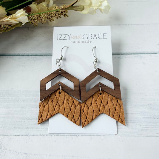 A pair of geometric dangle earrings displayed on a white card labeled 'IZZY and GRACE handmade.' The earrings have dark brown wood top sections and tan textured leather lower sections, hanging from silver hooks on a white wood plank background with dried flowers in the corner.