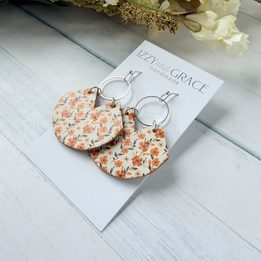 A pair of handmade semi-circular drop earrings with a warm-toned orange and brown floral pattern on a cream background, hanging from silver rings on a white display card. The card is branded 'IZZY and GRACE handmade' and is styled on a light gray wooden surface with dried flowers in the corner.