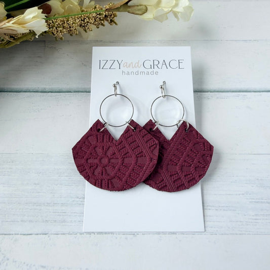 A pair of handmade semi-circular drop earrings in burgundy textured leather with silver-tone hoop connectors, displayed on a white card labeled 'IZZY and GRACE handmade' on a white wood background with dried flowers.