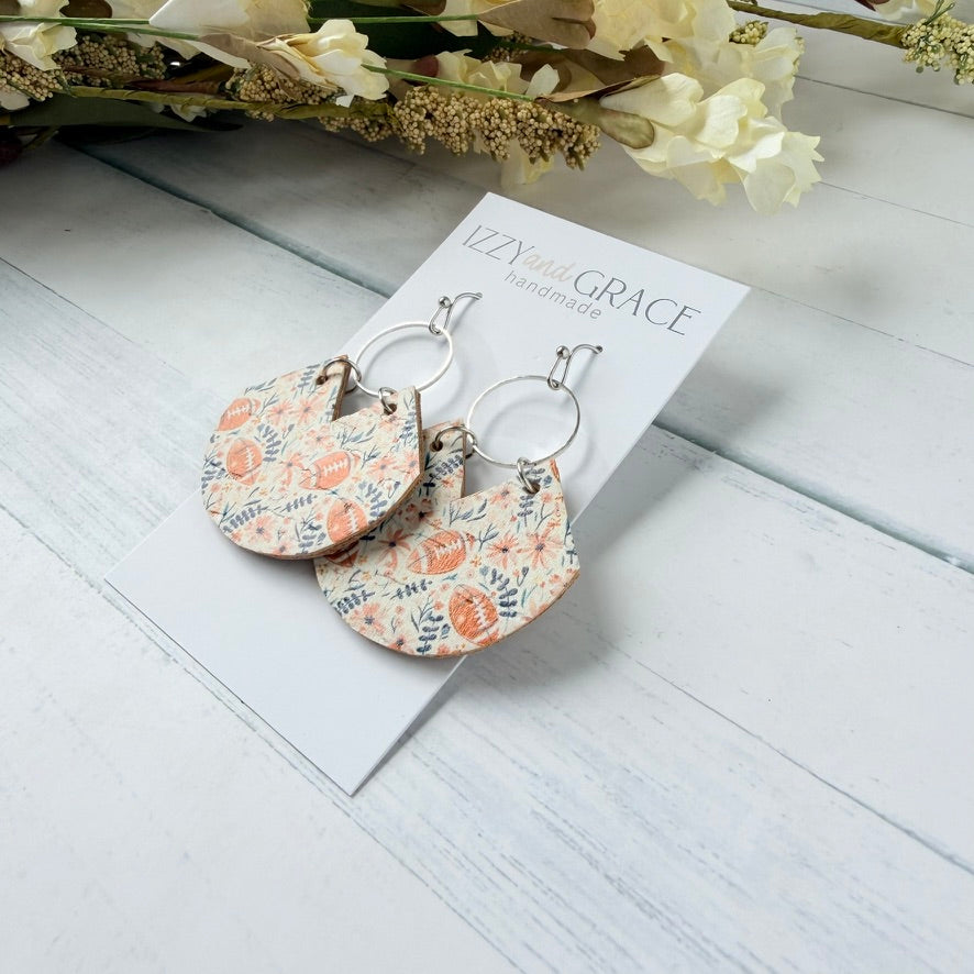 A pair of handmade dangle earrings with a floral patterned leather fan shape disc and silver hoop, displayed on a white card labeled 'Izzy's Grace Handmade' on a white wood background with dried flowers.