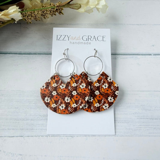 A pair of handmade dangle earrings with silver-tone hoop tops and semi-circular fabric drops featuring an orange, brown, and white floral pattern, displayed on a white card labeled 'IZZYand GRACE handmade' on a light wood background.