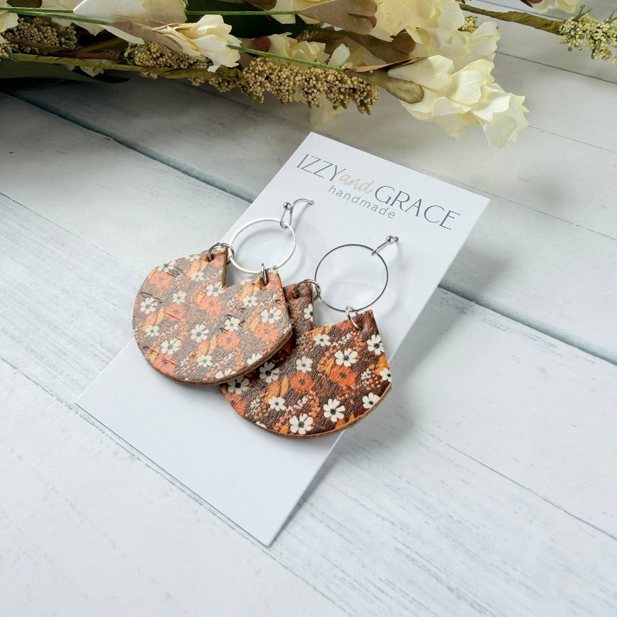 A pair of handmade semi-circular drop earrings with a brown, orange, and white floral pattern, hanging from silver rings on a white display card labeled 'Izzy's Grace Handmade'. The card is styled on a white wooden surface with dried flowers in the background.