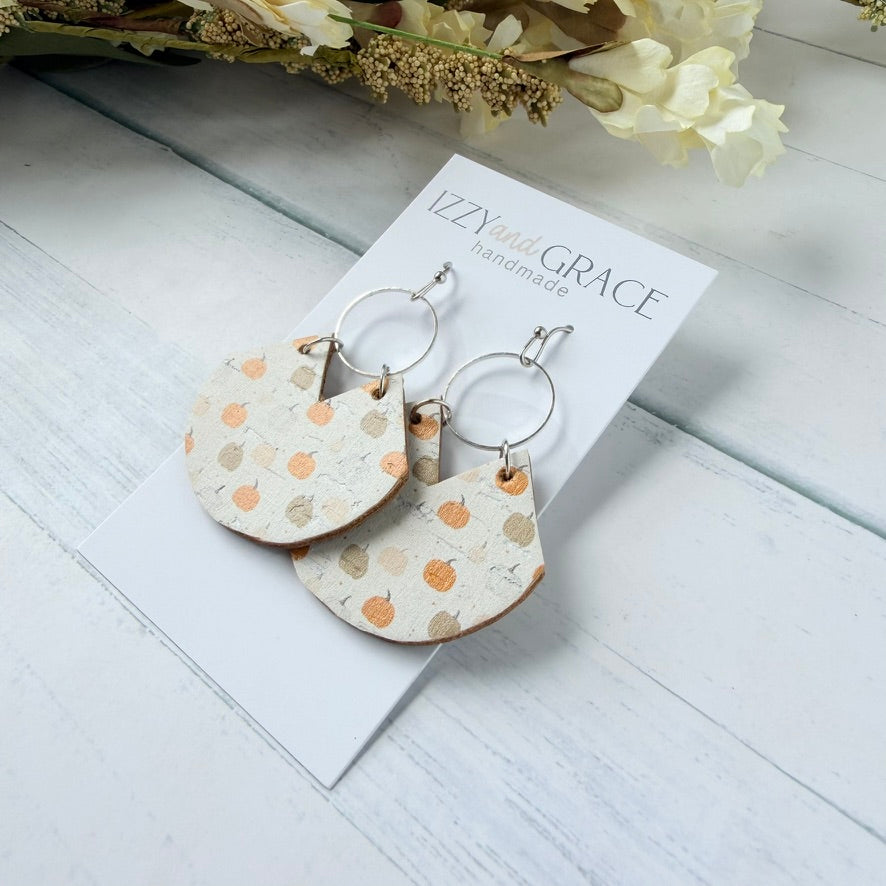 A pair of handmade dangle earrings displayed on a white card labeled 'Izzy and Grace handmade.' The earrings have silver-tone hoops and semi-circular beige charms decorated with a pattern of orange pumpkins and muted tan leaves.