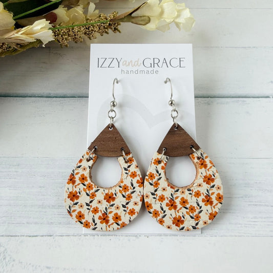 Teardrop-shaped dangle earrings with a cream background and orange floral print, attached to dark brown wooden triangles and silver hooks, displayed on a white card labeled 'IZZY and GRACE handmade' on a white wood surface.