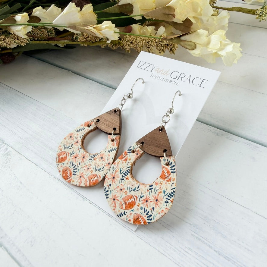 A pair of teardrop-shaped dangle earrings with a floral football pattern in pink and navy, attached to brown wooden triangles and silver hooks, displayed on a white card labeled 'Izzy and Grace handmade' on a white wood background with dried flowers.