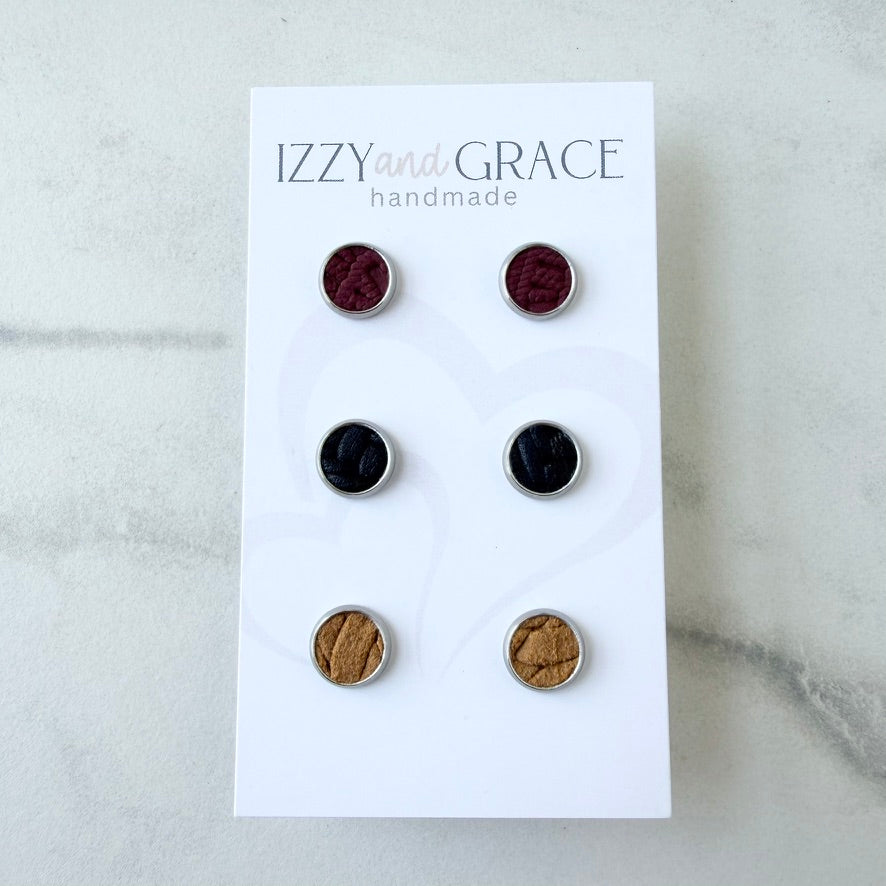 Six round stud earrings displayed on a white card labeled 'IZZY and GRACE handmade,' photographed on a light marble-like surface. The earrings come in burgundy, black, and tan/brown metallic finishes.