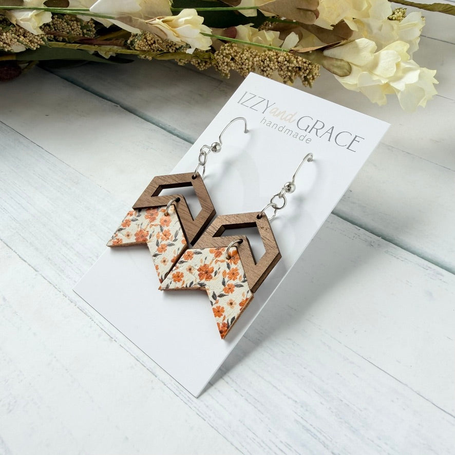 A pair of handmade geometric dangle earrings displayed on a white card labeled 'Izzy and Grace Handmade.' The earrings feature layered brown wood-like shapes over white leather with an orange floral print, hanging from silver hooks.