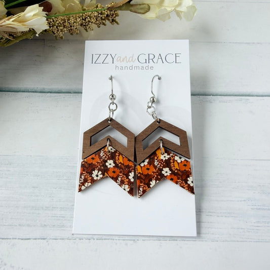 A pair of handmade geometric dangle earrings displayed on a white card labeled 'IZZY and GRACE handmade.' The earrings have brown wooden chevron shapes with orange, white, and black floral-patterned lower sections, hanging from silver fishhook wires.