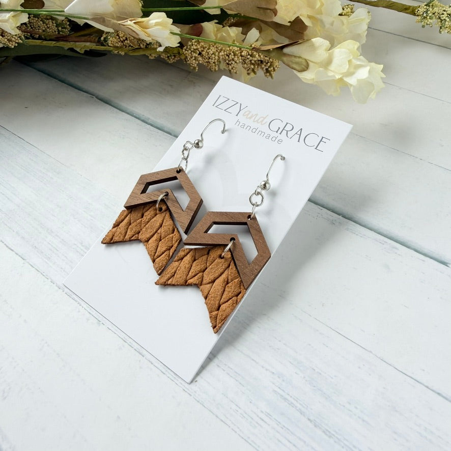 A pair of geometric drop earrings displayed on a white card labeled 'Izzy's Grace handmade.' The earrings have layered wood and leather chevron shapes in warm brown tones with silver fishhook wires, photographed on a white wood surface with dried flowers in the background.