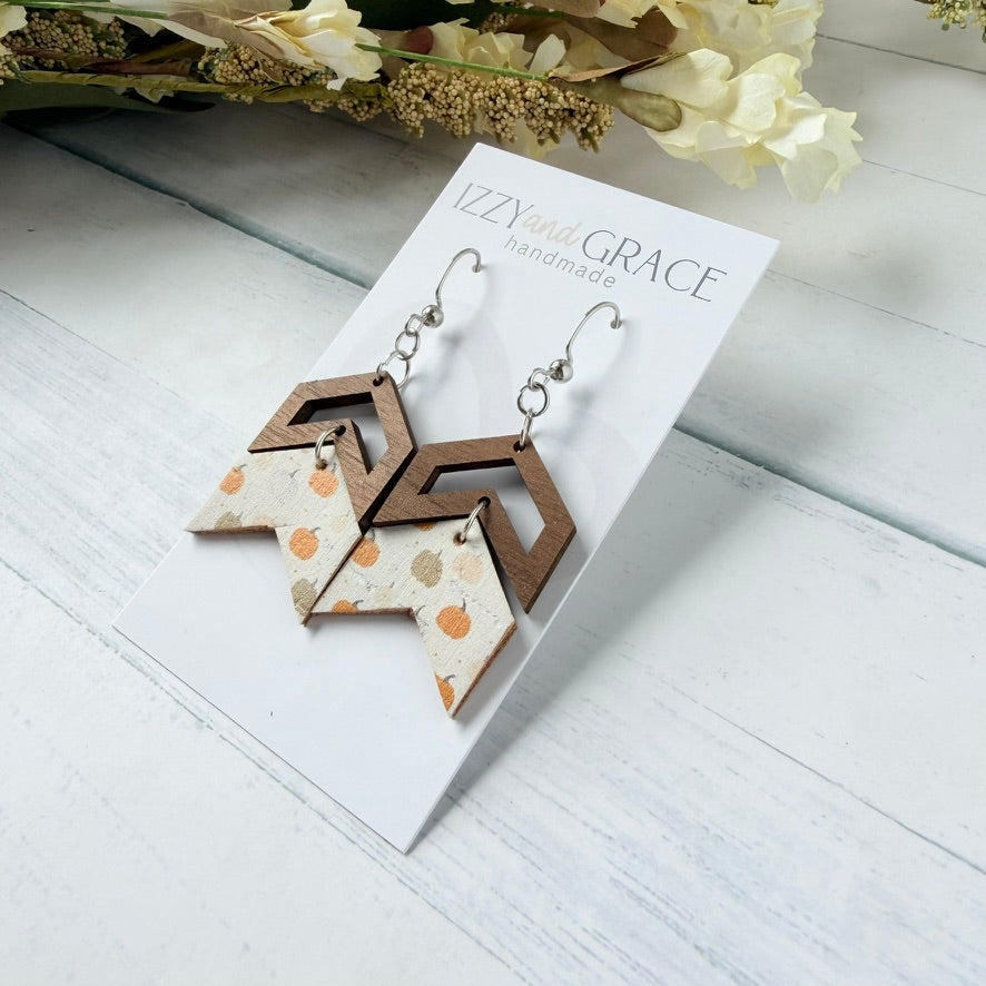 A pair of handmade geometric dangle earrings displayed on a white card labeled 'Izzy with Grace handmade.' The earrings have silver fishhook wires and chevron wooden tops in a medium brown, with leather bottoms with orange and tan pumpkins on a white background.