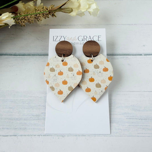 A pair of handmade dangle earrings displayed on a white card. The earrings feature a cream-colored leather petal-shaped drop with a pattern of orange and tan pumpkins, attached to a round dark brown wooden stud.