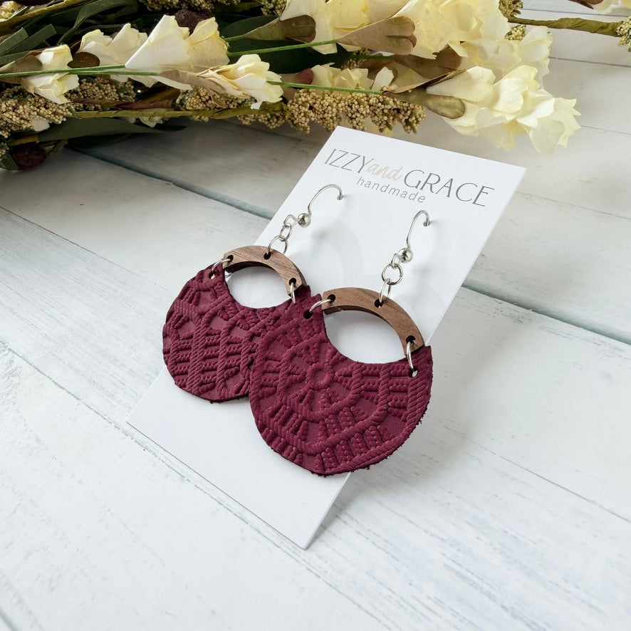 A pair of handmade dangle earrings displayed on a white card. The earrings feature textured burgundy circular drop pieces with a crochet-like pattern, connected by a wood arch and hanging from silver-toned hooks.