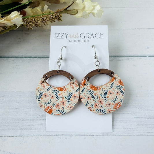 A pair of handmade dangle earrings displayed on a white card labeled 'IZZY and GRACE handmade'. The earrings feature circular leather with a floral pattern in orange, blue, white, and footballs connected by brown wooden arches and hanging from silver fishhook wires.
