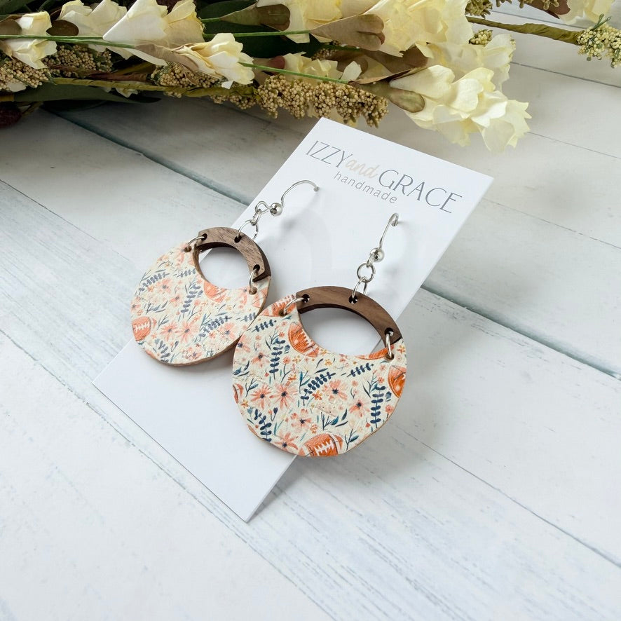A pair of handmade dangle earrings displayed on a white card labeled 'Izzy and Grace Handmade.' The earrings have a floral patterned disc with pink and blue botanical football motifs, a brown wooden arch, and silver-tone hooks.