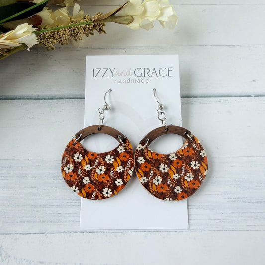 A pair of round drop earrings with a brown and orange floral pattern, displayed on a white card labeled 'IZZY and GRACE handmade' on a white wood background with dried flowers.