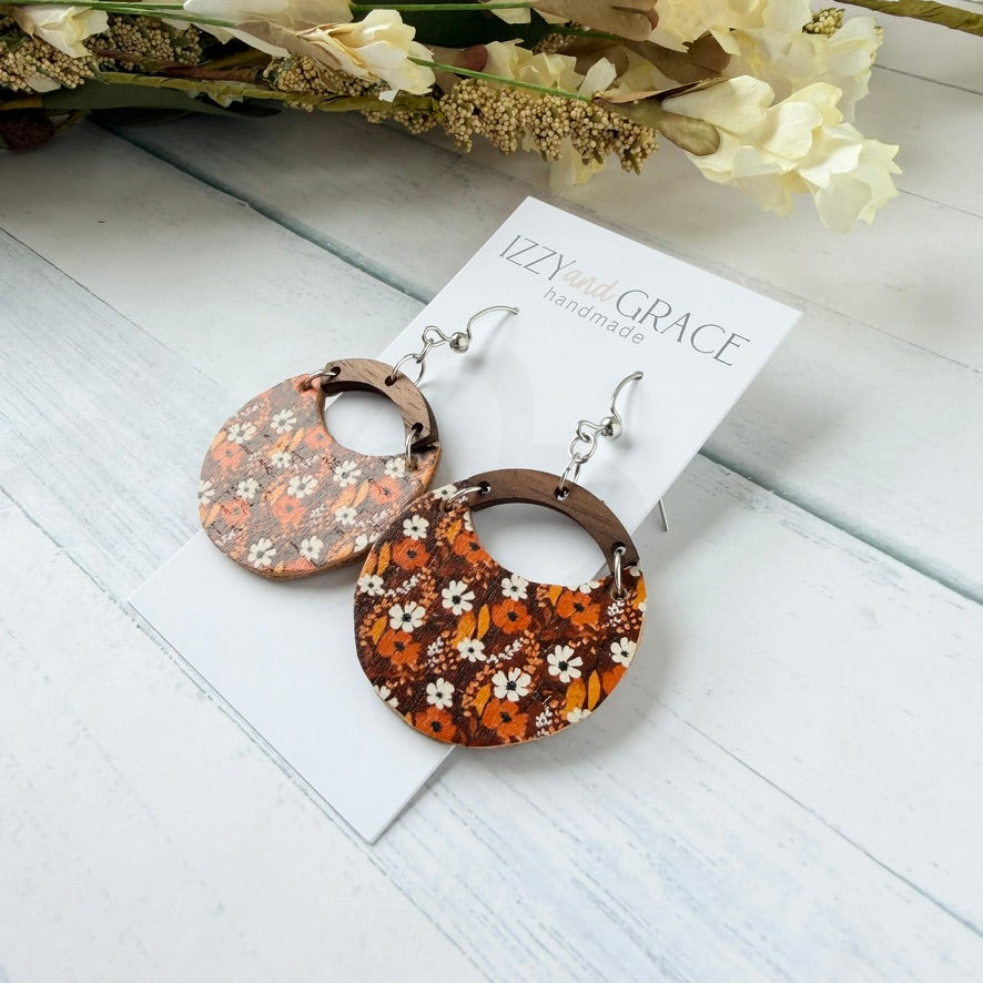 A pair of round drop earrings with a brown, orange, and white floral pattern, attached to silver hooks and displayed on a white card labeled 'IZZY and GRACE handmade' on a white wood background.