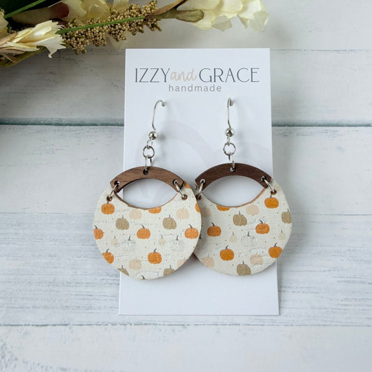A pair of handmade dangle earrings displayed on a white card labeled 'IZZY and GRACE handmade.' The earrings have silver fishhook wires and circular beige leather bottom with an orange and tan pumpkin pattern, connected by brown wooden arches.