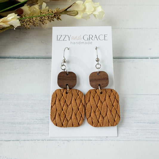 A pair of wooden drop earrings with textured mustard leather lower sections and darker brown wooden square connectors, displayed on a white card labeled 'IZZY and GRACE handmade' on a whitewashed wood surface.