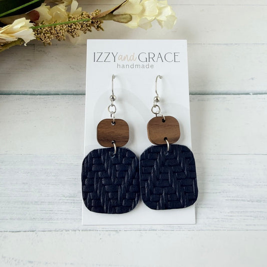 A pair of handmade dangle earrings displayed on a white card. Each earring has a silver fishhook wire, a small rounded square wooden top, and a larger rectangular navy leather bottom.