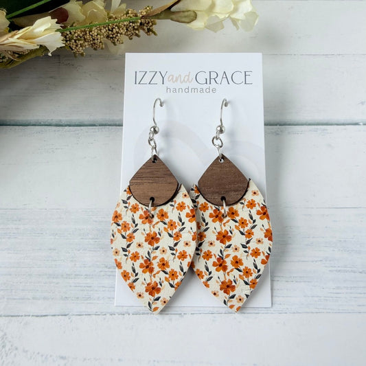 A pair of handmade dangle earrings displayed on a white card labeled 'IZZY and GRACE handmade.' The earrings have silver fishhook wires, brown wooden teardrop tops, and cream leather bottoms with an orange floral print.