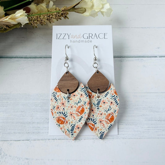 A pair of handmade petal-shaped dangle earrings displayed on a white card labeled 'IZZY and GRACE handmade.' The earrings have silver fishhook wires, medium brown wooden teardrop tops, and lower leather panels with a floral and football print in pink, navy, and cream.
