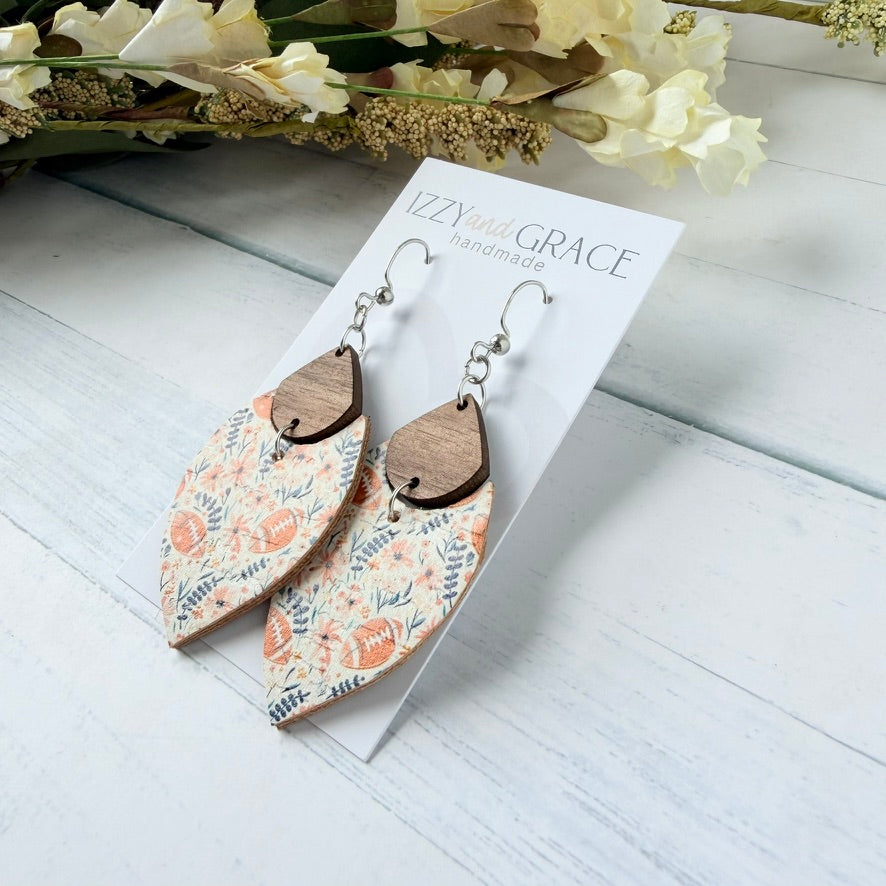 A pair of handmade dangle earrings displayed on a white card labeled 'IZZY and GRACE handmade'. The earrings feature a floral patterned leather bottom with pink flowers, blue leaves, and footballs, attached to a small brown wooden teardrop tup and silver fishhook wires.