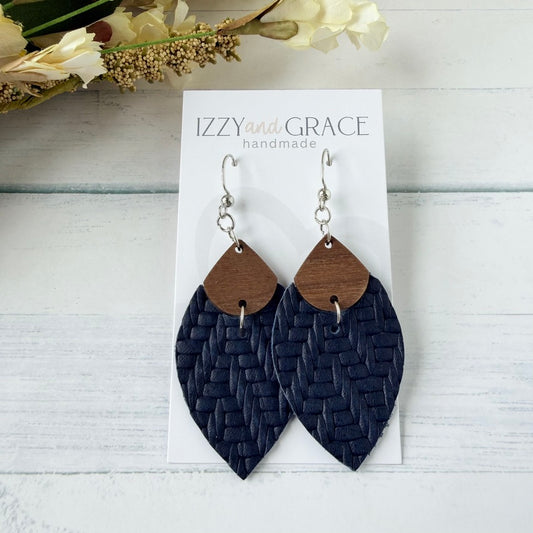 A pair of handmade dangle earrings displayed on a white card labeled 'IZZYand GRACE handmade.' The earrings have silver-tone hooks, small brown wooden teardrop shapes, and larger woven navy blue petal-shaped drops.