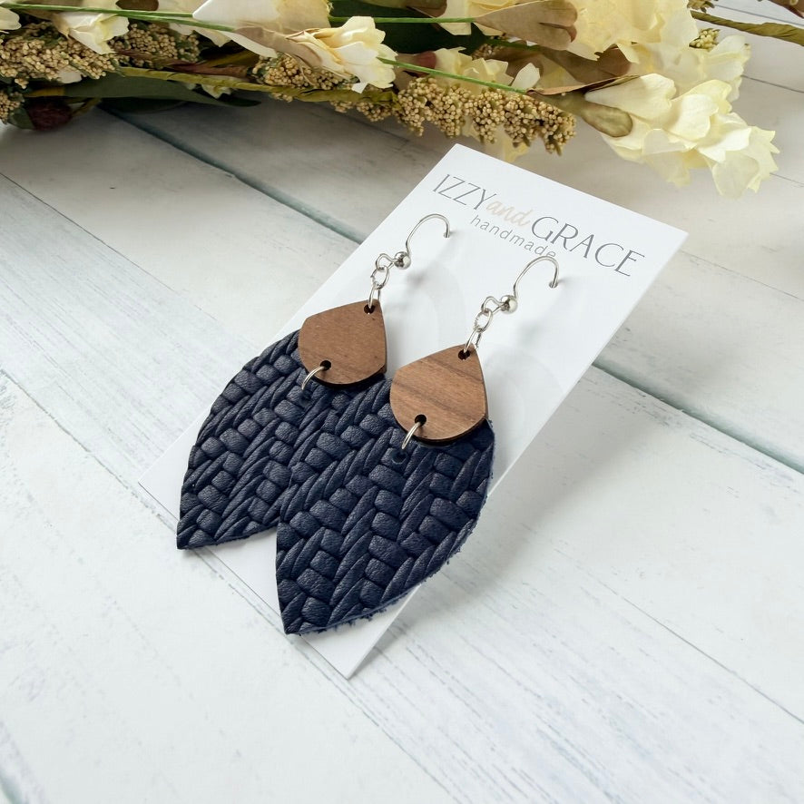 A pair of handmade dangle earrings displayed on a white card. The earrings feature dark navy blue woven leather petal-shaped drops with small natural wood accents and silver-tone hooks.