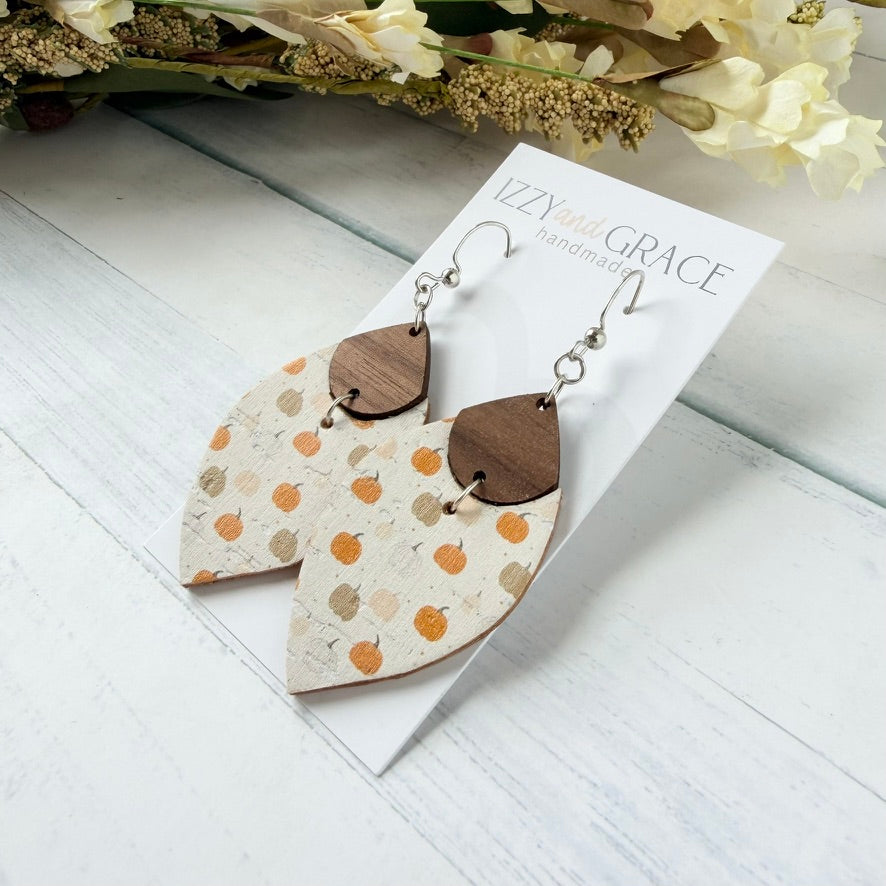 A pair of handmade dangle earrings displayed on a white card labeled 'Izzy and Grace Handmade.' The earrings have petal-shaped beige and white wood pieces with orange and tan pumpkin motifs, connected by small brown wooden teardrops and silver fishhook ear wires.