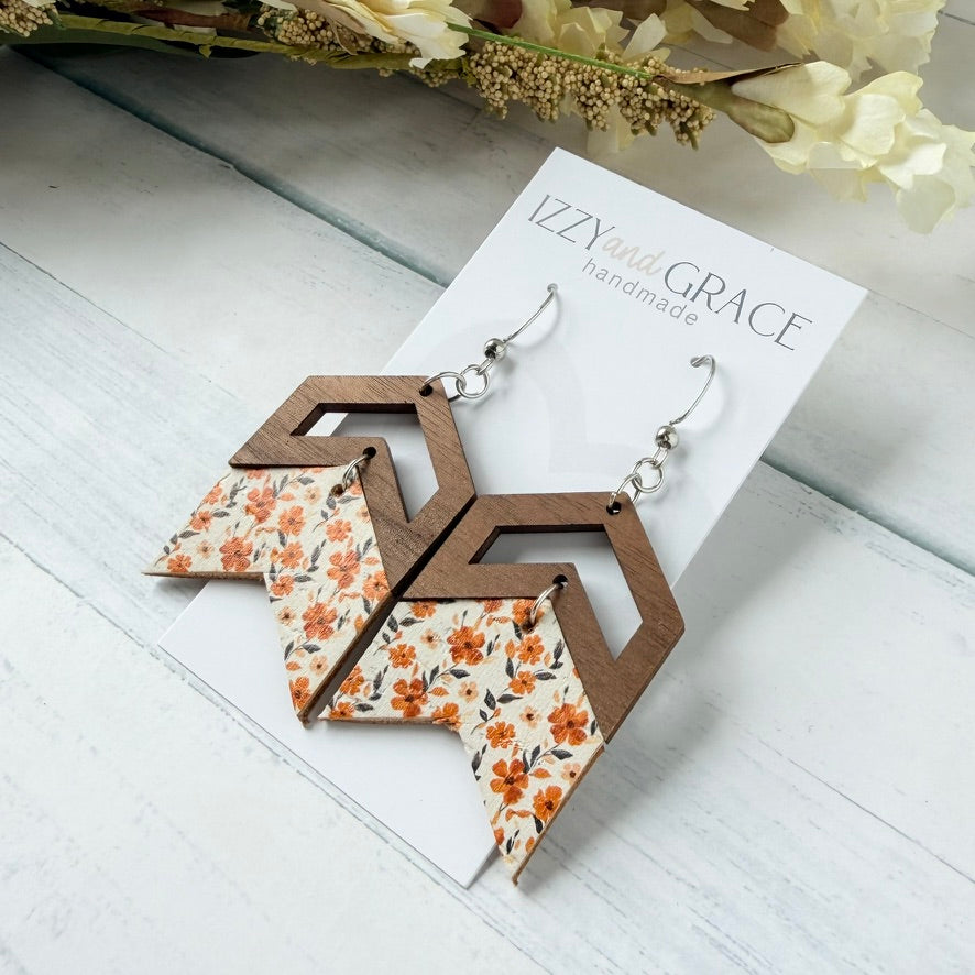 A pair of geometric drop earrings displayed on a white card labeled 'IZZY and GRACE handmade.' The earrings have dark brown wooden chevron tops, silver hooks, and lower panels with an orange, beige, and gray floral pattern.