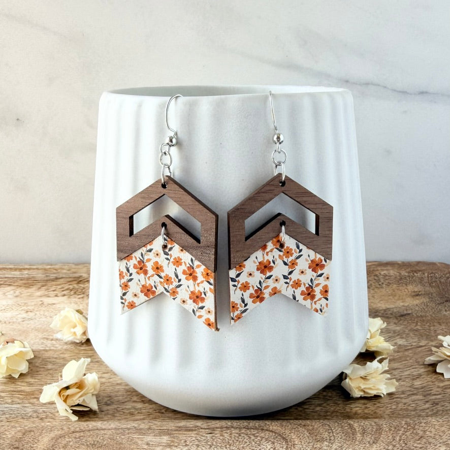 A pair of geometric drop earrings displayed on a white ribbed ceramic cup. The earrings feature a dark brown wooden chevron top and a white fabric chevron bottom printed with an orange and green floral pattern.