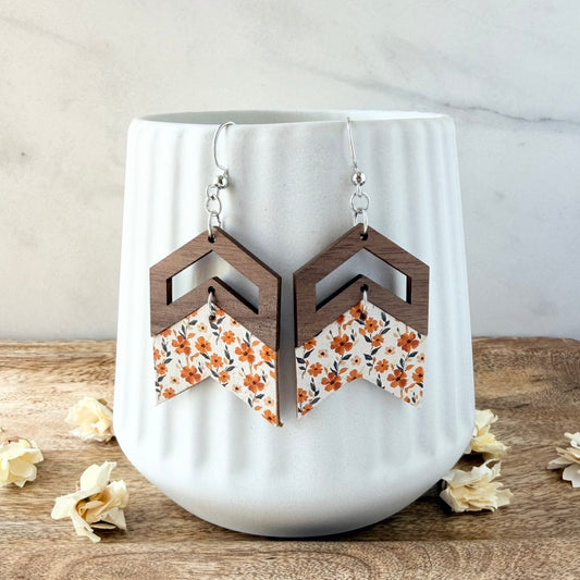 A pair of geometric drop earrings displayed on a white ribbed ceramic cup. The earrings feature a dark brown wooden chevron top and a white fabric chevron bottom printed with an orange and green floral pattern.