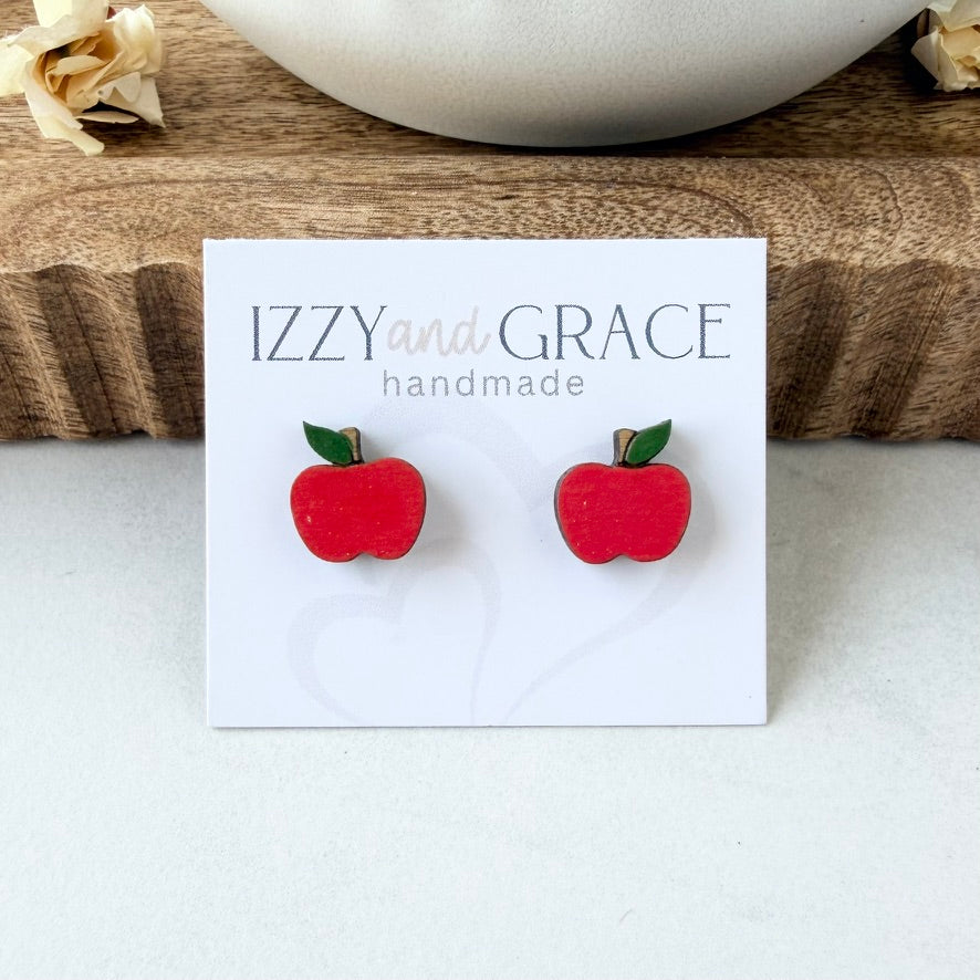 A pair of red apple stud earrings with green leaves on a white display card that reads 'IZZY and GRACE handmade', photographed on a light surface with a wooden tray and white bowl in the background.