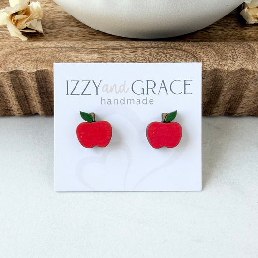 A pair of red apple stud earrings with green leaves on a white display card that reads 'IZZY and GRACE handmade', photographed on a light surface with a wooden tray and white bowl in the background.