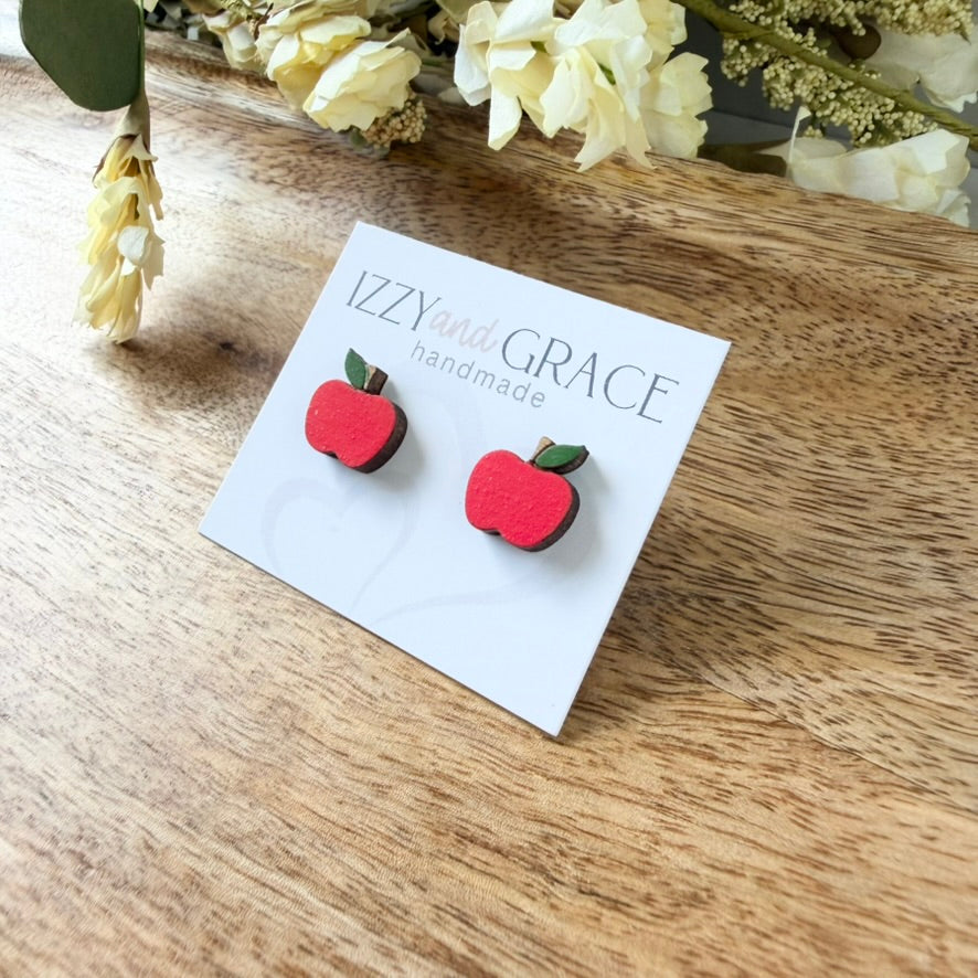 A pair of red apple stud earrings with green leaves on a white display card that reads 'IZZYand GRACE handmade', placed on a wooden surface with dried flowers in the background.