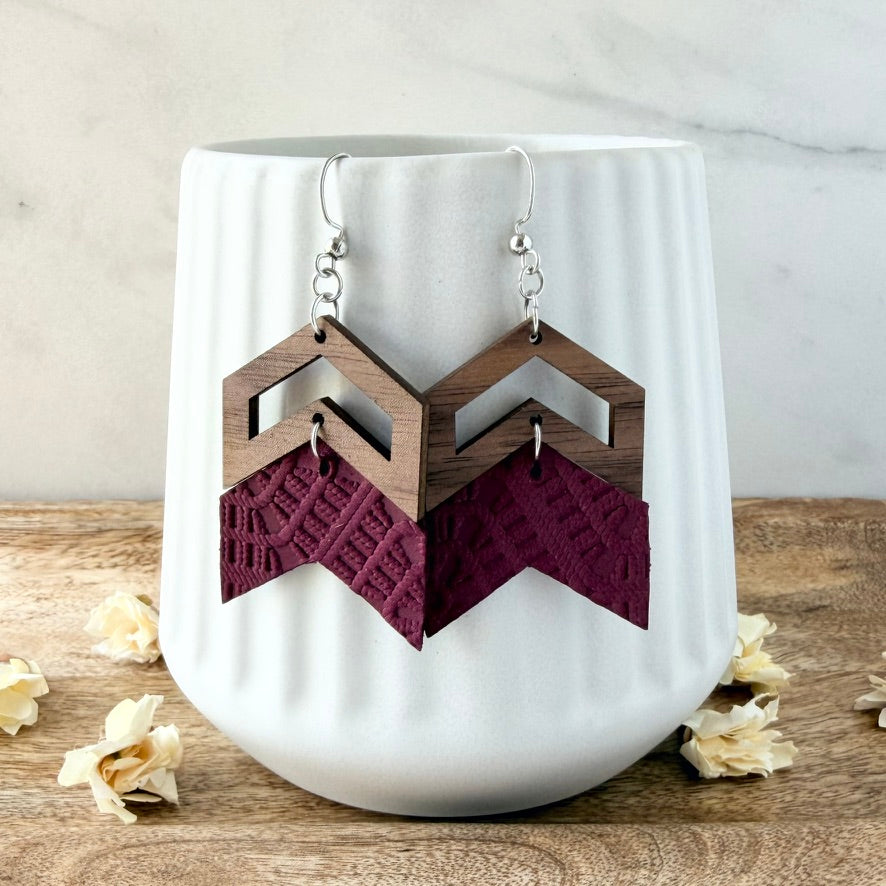A pair of geometric drop earrings with dark brown wood and deep burgundy textured fabric, hanging from silver fishhook wires in front of a white ribbed ceramic cup on a wooden surface.