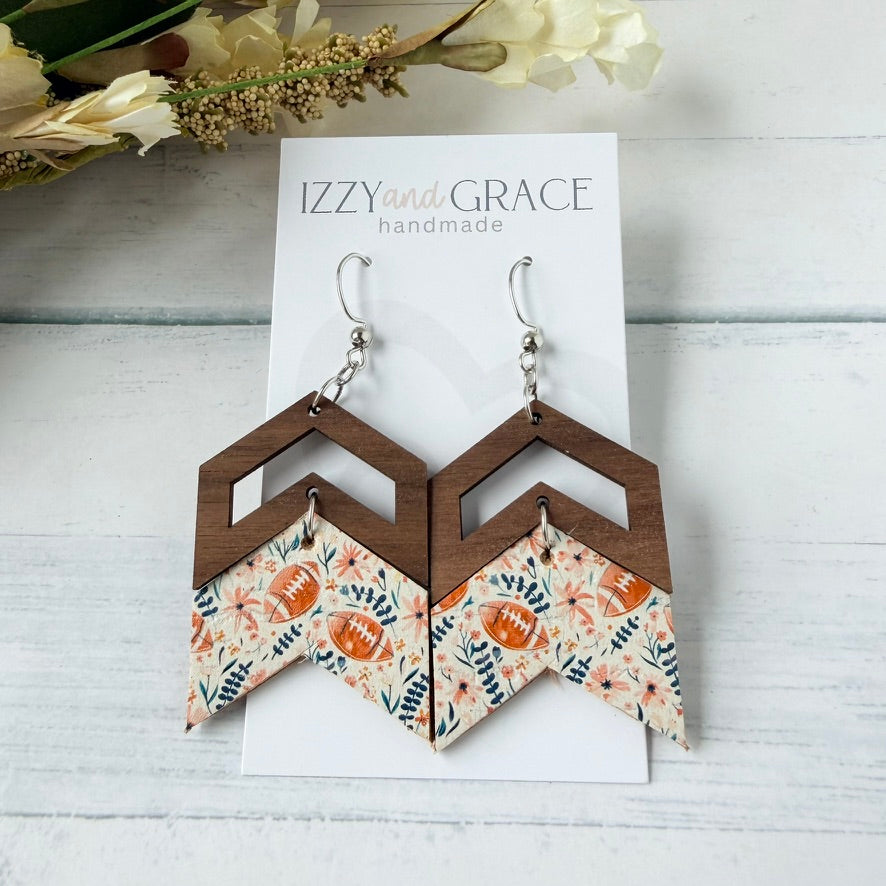 A pair of handmade geometric dangle earrings displayed on a white card labeled 'IZZY and GRACE handmade.' The earrings have a dark brown wooden top section and a lower chevron section with a floral print featuring orange football motifs.