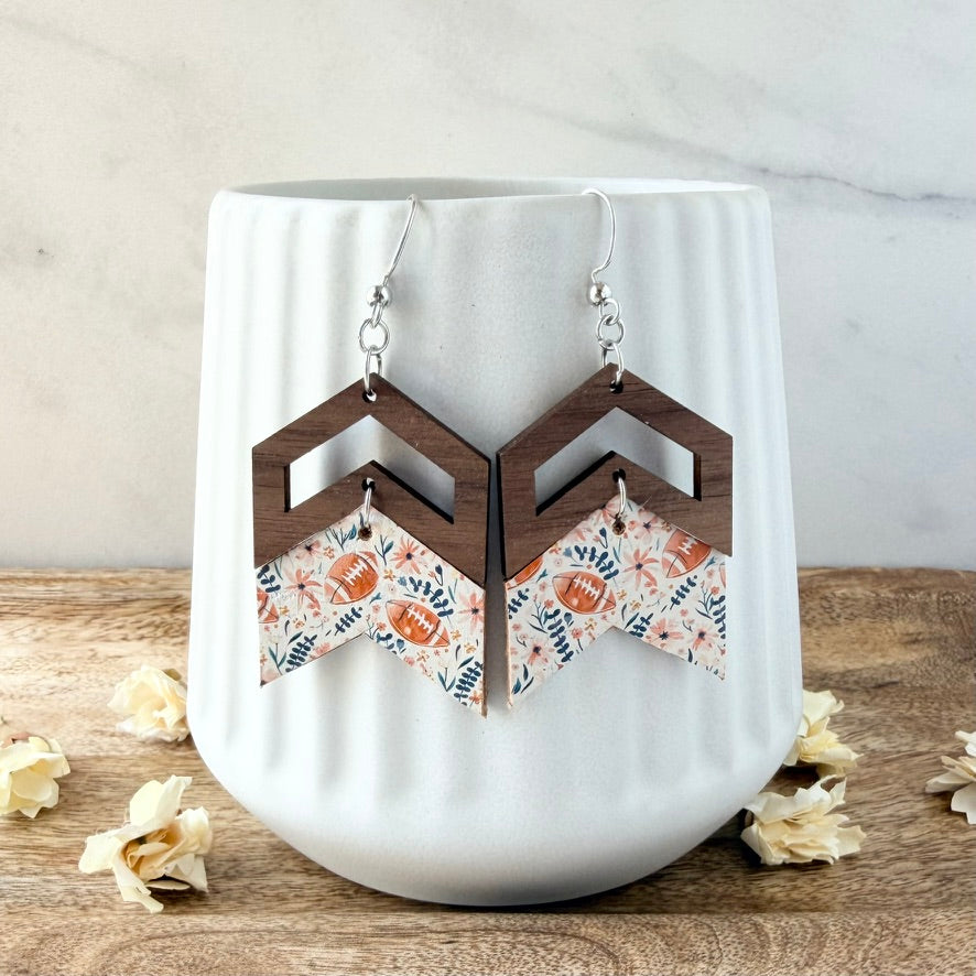 A pair of geometric drop earrings displayed on a white ribbed ceramic cup. The earrings feature dark brown wooden chevron frames with patterned triangular dangles in a floral and football print on a white background, resting on a wooden surface with scattered popcorn kernels.
