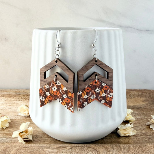 A pair of geometric drop earrings displayed on a white ribbed ceramic vase. The earrings feature a dark brown wooden geometric top with a chevron-shaped lower flap decorated with an orange, brown, and white floral pattern.