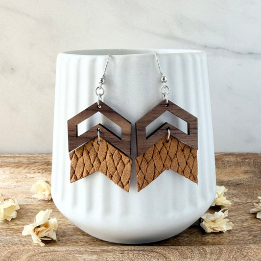 A pair of geometric dangle earrings with dark brown wood top halves and textured tan leather bottom halves, hanging on a white ribbed ceramic cup on a wooden surface with dried flowers.