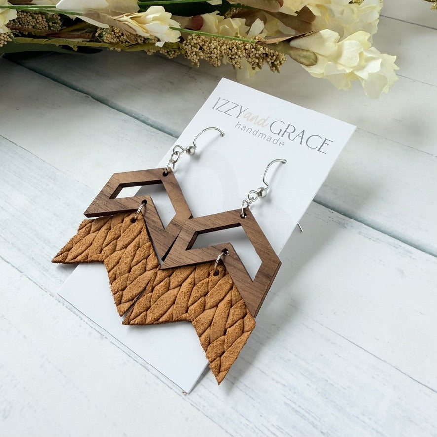 Wood and leather geometric drop earrings displayed on a white card labeled 'Izzie and Grace handmade' on a white wood surface with dried flowers in the background.
