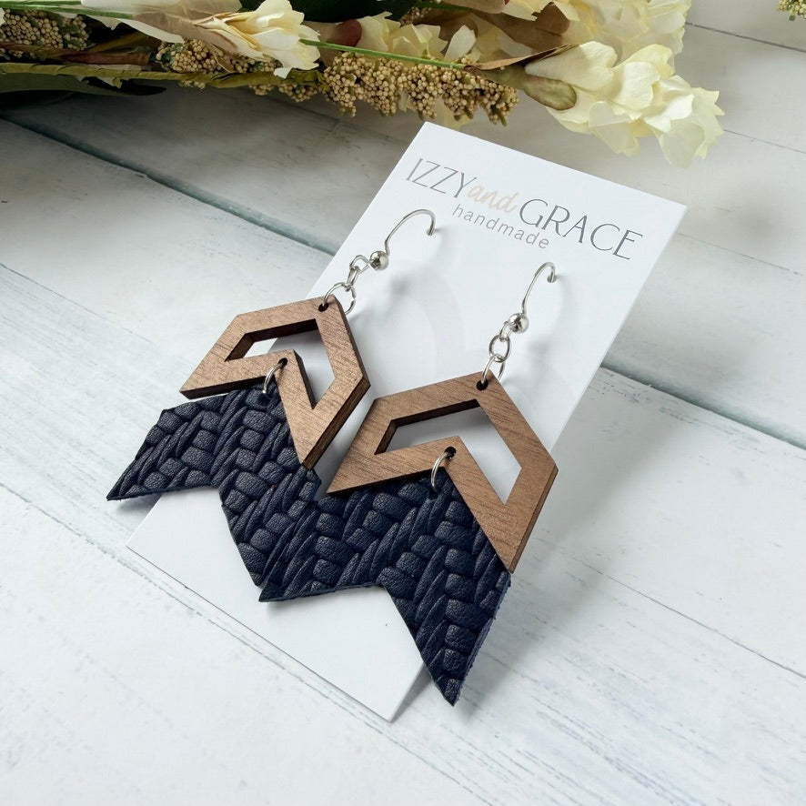 A pair of handmade geometric dangle earrings displayed on a white card. The earrings feature a dark navy blue woven leather lower section and a light brown wood upper section with silver-tone hooks.