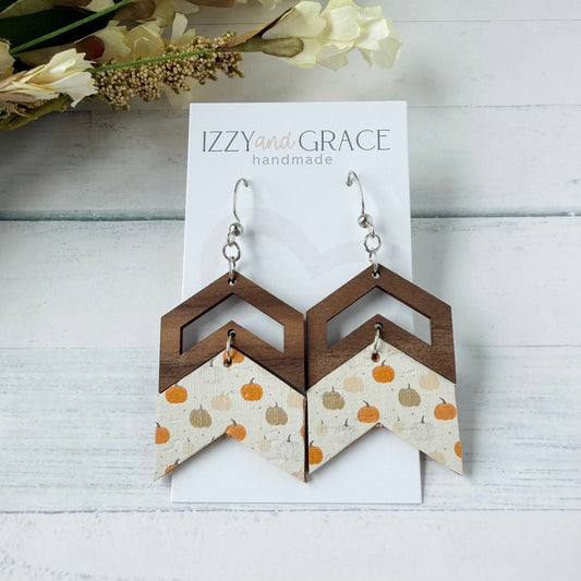 A pair of handmade geometric dangle earrings displayed on a white card labeled 'IZZY and GRACE handmade.' The earrings have dark brown wooden chevron tops, silver hooks, and lower panels with a cream background and orange, peach, and tan pumpkin motifs.
