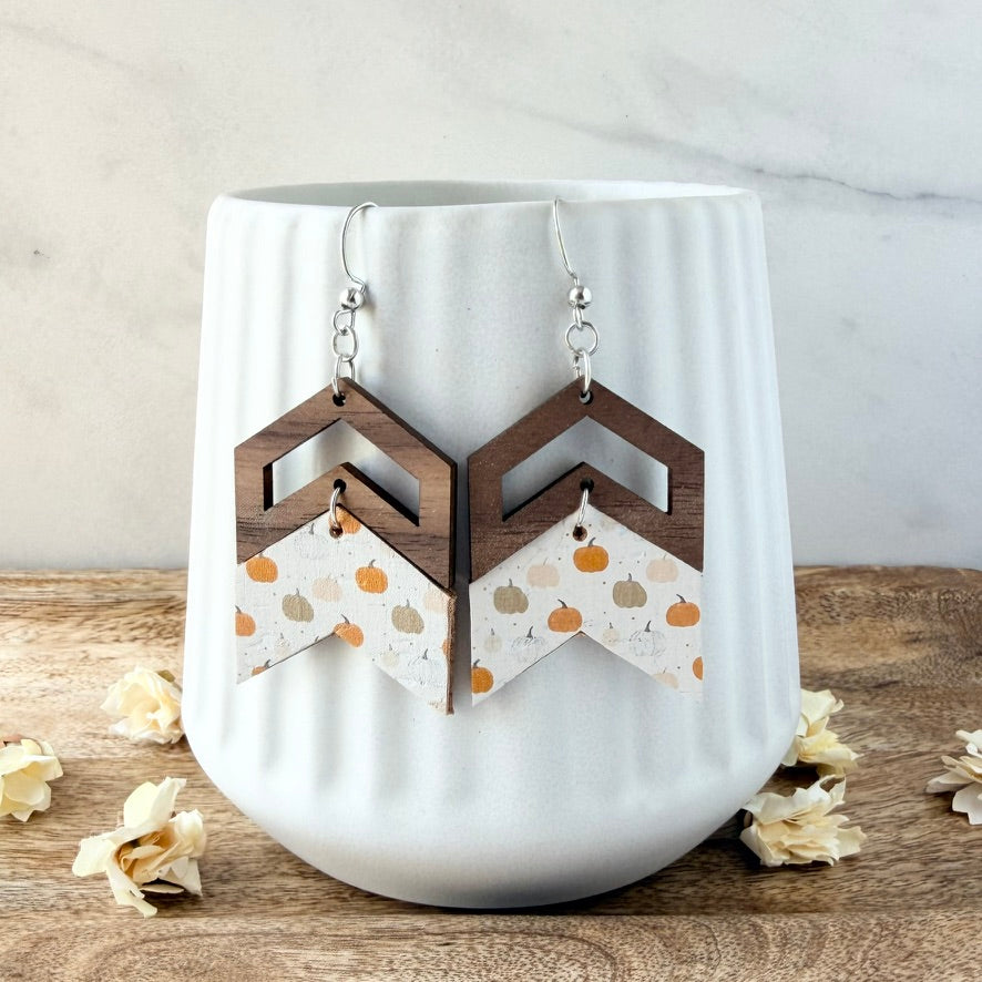 A pair of geometric drop earrings with dark brown wooden chevron tops and white paper dangles printed with orange pumpkins and small tan leaves, hanging in front of a white ribbed ceramic cup on a wooden surface with dried flowers.