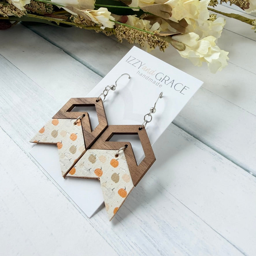 A pair of handmade geometric dangle earrings displayed on a white card labeled 'IZZY and GRACE handmade.' The earrings have dark brown wooden chevron tops, silver hooks, and lower panels with a cream background and orange, peach, and tan pumpkin motifs.