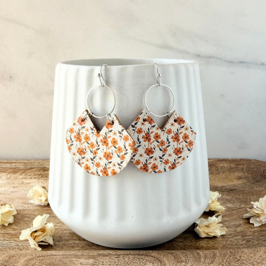A pair of handmade dangle earrings with orange and brown floral fabric charms, displayed on a white ribbed ceramic cup on a wooden surface.