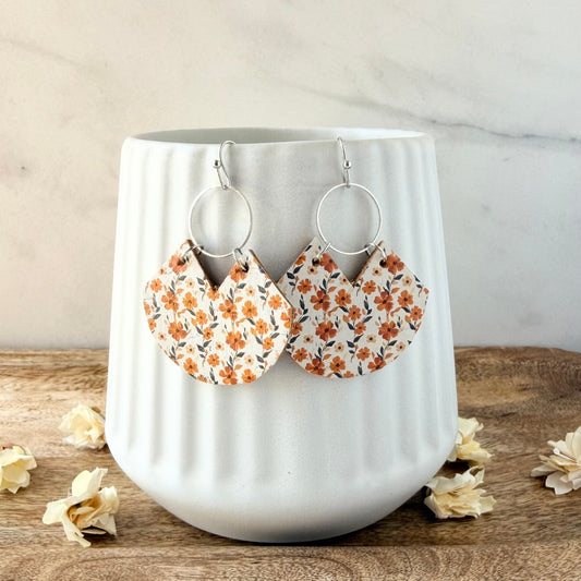 A pair of handmade dangle earrings with orange and brown floral fabric charms, displayed on a white ribbed ceramic cup on a wooden surface.