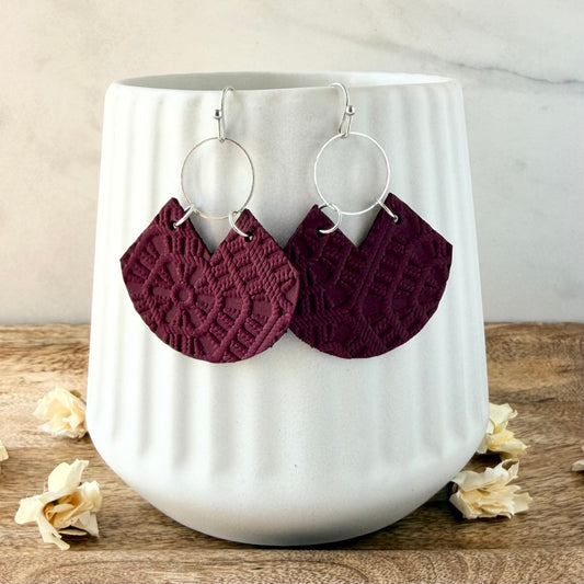 A pair of handmade dangle earrings with silver-tone circular hoops and textured, patterned burgundy fan shapes, displayed on a white ribbed ceramic mug.