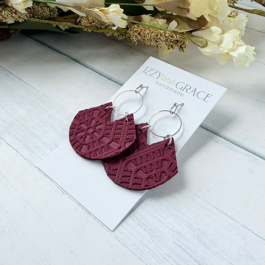 A pair of handmade dangle earrings with textured burgundy semicircle drops and silver-tone hoops, displayed on a white card labeled 'IZZYBEAR & GRACE handmade' on a white wood background with dried flowers.